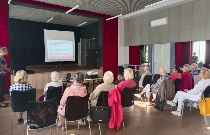 Zdjęcie przedstawia uczestników warsztatu pod tytułem Profilaktyka odleżyn, którzy słuchają wykładu pielęgniarki i oglądają kolejny slajd prezentacji tematycznej. Seniorzy siedzą na krzesłach przed sceną w głównej sali Centrum Aktywności Seniora.