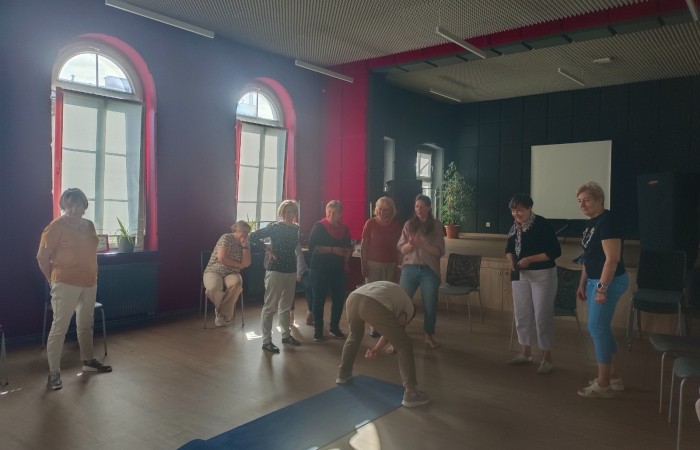 Zdjęcie przedstawia uczestników warsztatu pod tytułem Terapia śmiechem zebranych w sali Centrum Aktywności Seniora. Grupa ośmiu seniorek i instruktorka patrzą na jedną z uczestniczek, która wykonuje śmiesznie wyglądającą figurę.