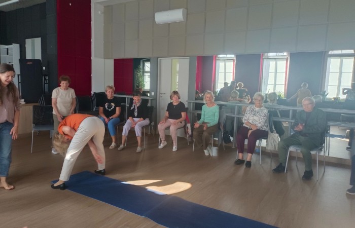 Zdjęcie przedstawia uczestników warsztatu pod tytułem Terapia śmiechem siedzących na krzesłach w sali Centrum Aktywności Seniora. Uśmiechnięci seniorzy i instruktorka patrzą na jedną z uczestniczek, która wykonuje śmiesznie wyglądającą figurę.