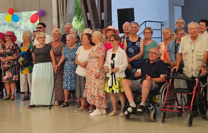 Na zdjęciu widać grupę seniorów w towarzystwie organizatorów, którzy pozują do zdjęcia rozpoczynającego Letnią dyskotekę dla Seniorów. Miejsce, w którym rozgrywa się wydarzenie, jest przystrojone kolorowymi ozdobami.