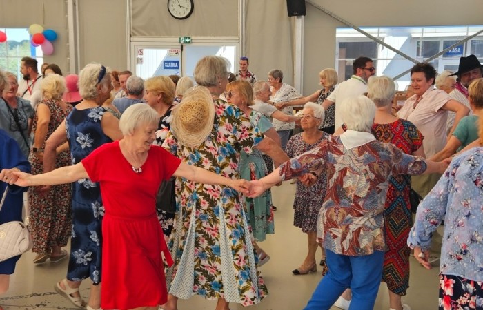 Zdjęcie przedstawia kolorowo ubranych seniorów, którzy tańczą połączeni w jeden długi łańcuch