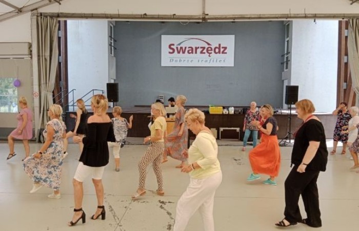 Fotografia ukazuje kobiety, które tańczą taniec Jerusalema. Uchwycono moment, kiedy dotykają w rytmie muzyki górną częścią stopy podłoże. W tle widać 
DJ-a oraz wodzireja imprezy.
