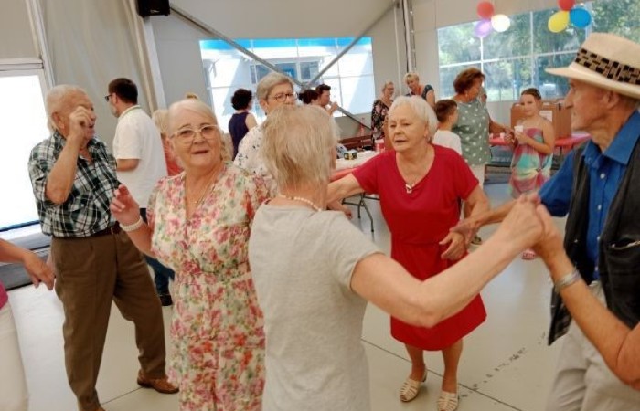 Na pierwszym planie zdjęcia widać bawiąca się w rytm muzyki grupę seniorów. W tle widać organizatorów, którzy wydają uczestnikom ciasto oraz kawę i herbatę. 