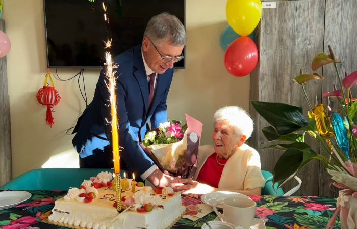 Uroczystość sto trzecich urodzin Pani Franciszki Nowak w Dziennym Domu Pomocy. Sala jest skąpana w słońcu, ozdobiona kolorowymi balonami, kwiatami i papierowymi dekoracjami wykonanymi ręcznie przez uczestników i kadrę placówki. Na pierwszym planie burmistrz miasta i gminy Swarzędz – pan Marian Szkudlarek wręcza bukiet kwiatów Jubilatce. Przed Panią Franciszką na stole stoi tort z zapalonymi świeczkami oraz racą, z której wylatuje strumień iskier. Jubilatka i jej gość są elegancko ubrani i mają roześmiane twarze.