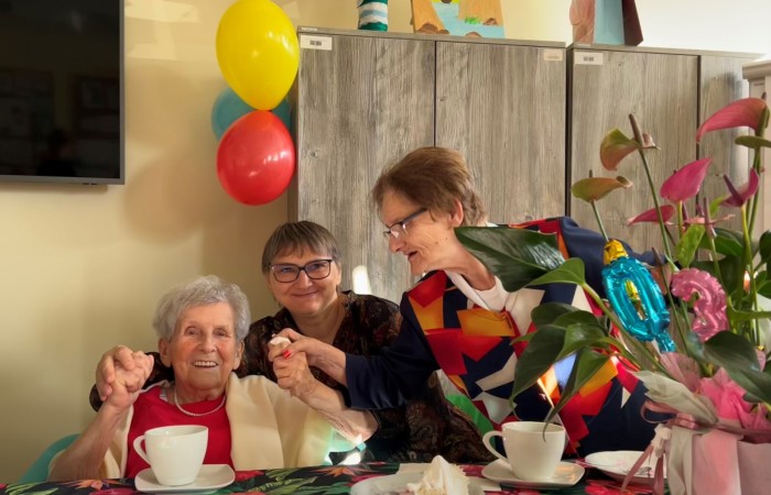 Za urodzinowym stołem w Dziennym Domu Pomocy siedzi uradowana stutrzylatka, którą od tyłu trzyma za dłonie uśmiechnięta opiekunka Eugenia Zielke. Od prawej pochyla się nad nimi pani Asia, uczestniczka zajęć. Kobiety trzymają się za ręce i podrygują do muzyki. Przed nimi na stole stoją kubki, talerze z tortem oraz kolorowo przystrojona doniczka z anturium.