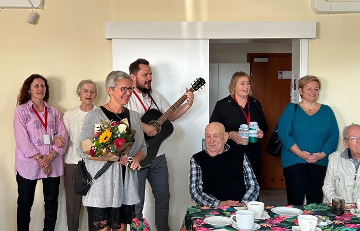 Uroczystość sto trzecich urodzin Pani Franciszki Nowak w Dziennym Domu Pomocy. Roześmiani goście śpiewają 200 lat Jubilatce. Wśród osób widocznych na zdjęciu są dyrektor Centrum Usług Społecznych, Agnieszka Maciejowicz i zastępca dyrektora Centrum Usług Społecznych w Swarzędzu, Jolanta Osada, pracownicy i seniorzy z Dziennego Domu Pomocy. W grupie gości wyróżnia się jeden z pracowników, Rafał Wójtowicz, który gra na gitarze i śpiewa.