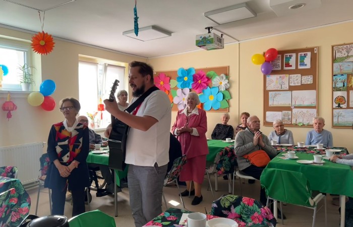 Wnętrze Sali w Dziennym Domu Pomocy, oświetlonej słońcem, udekorowanej ręcznie zrobionymi ozdobami. Seniorzy – uczestnicy zajęć, pod wodzą pracownika Rafała Wójtowicza grającego na gitarze, śpiewają Jubilatce 200 lat i klaszczą do rytmu. Część z nich stoi, część siedzi za stołami nakrytymi zielonym obrusem i zastawionymi filiżankami z kawą i herbatą.