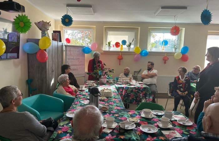 Urodziny Pani Franciszki Nowak w Dziennym Domu Pomocy. W głębi sali po lewej stronie stoi tablica, a na niej zawieszony jest plakat z napisem „Sto trzecie urodziny Pani Frani”. Stoły w sali zostały ustawione w kształcie litery ce, przykryte są obrusami ze wzorem egzotycznych różnokolorowych kwiatów. Na stole stoją: tort urodzinowy, termos, białe talerzyki, białe filiżanki z kawą i herbatą oraz udekorowana doniczka z anturium. Zgromadzeni, a wśród nich Jubilatka siedzą przy stole i zwróceni są w kierunku przemawiającej w głębi sali pani Stanisławy Krzemieniewskiej, byłej prezes Związku Emerytów, Rencistów i Inwalidów.