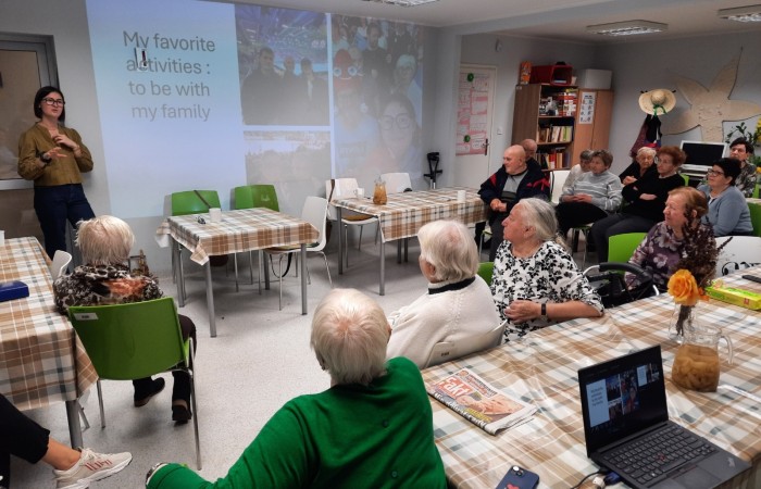 Wnętrze głównej sali w Dziennym Domu Senior Wigor przy ulicy Sienkiewicza 21 w Swarzędzu. Wolontariuszka Europejskiego Korpusu Solidarności Żuliet Żama pokazuje swoją pierwszą prezentację. Na ścianie widać slajd poświęcony ulubionym aktywnościom Żuliet, a dokładnie spędzaniu czasu ze swoją rodziną. Slajd składa się z trzech zdjęć. Pierwsze przedstawia Żuliet z bratem Batist i dziadkiem Lui na meczu piłki ręcznej. Drugie zdjęcie przedstawia Żuliet z tatą Pier-iw na trybunach wśród kibiców na Paraolimpiadzie w Paryżu. Trzecia fotografia przedstawia Żuliet z dziadkiem Lui na meczu koszykówki w Szolet. Dookoła sali stoją stoły przybrane ceratą. Przed stołami na zielonych krzesłach siedzi trzynaścioro seniorów. Wszyscy są zasłuchani i patrzą na stojącą przy ścianie Żuliet, która opowiada o swojej rodzinie.