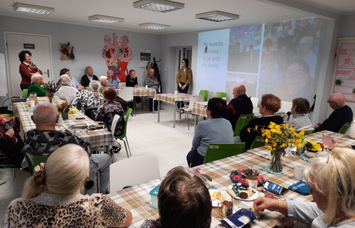 Wnętrze głównej sali w Dziennym Domu Senior Wigor. Na ścianie jest wyświetlony slajd z ulubionym zajęciem wolontariuszki Żuliet, czyli spędzaniem czasu z najbliższymi. W centrum kadru stoi Żuliet, dookoła niej i przed nią siedzą seniorki, seniorzy i członkowie kadry. Uwagę zwracają stoliki ubrane w ceratę w kratkę, na których w różnych miejscach stoją wazony z kwiatami. W lewym górnym rogu zdjęcia za stołem, przy którym siedzą seniorzy, stoi kierownik placówki, Anna Szpakowska, która tłumaczy seniorom jedną z wypowiedzi Żuliet.