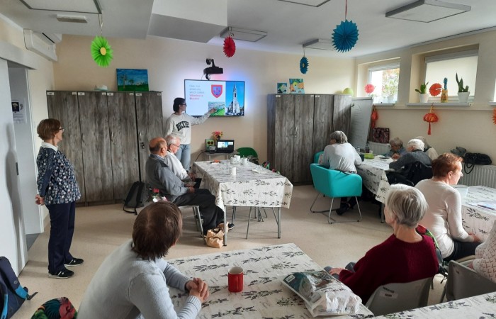 Wnętrze głównej sali w Dziennym Domu Pomocy przy ulicy Sienkiewicza 21. Przy stołach przybranych w ceraty z motywem gałązek lawendy siedzą seniorki i seniorzy - uczestnicy zajęć. Po lewej stronie przy wejściu do sali stoi jedna z uczestniczek, pani Asia. Wszyscy są zwróceni w kierunku wolontariuszki Żuliet Żama stojącej przy ścianie, która pokazuje na ekranie slajd ze swojej pierwszej prezentacji. Wolontariuszka wskazuje palcem zdjęcie przedstawiające widok miasteczka, z którego pochodzi - Wildju la Bluer. Na drugim zdjęciu jest widoczny stary kościół z wysoką i strzelistą wieżą - główna świątynia Wildju.