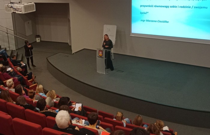 Sala wykładowa z publicznością. Na scenie kobieta w ciemnym stroju przemawia do mikrofonu, stojąc za przezroczystą mównicą z herbem miasta Swarzędz. Na ekranie wyświetlany jest temat prezentacji: „Kiedy życie traci barwy... Psychoterapia, czyli jak przywrócić równowagę sobie i rodzinie/swojemu życiu?”. W tle tablica z napisem "JA".