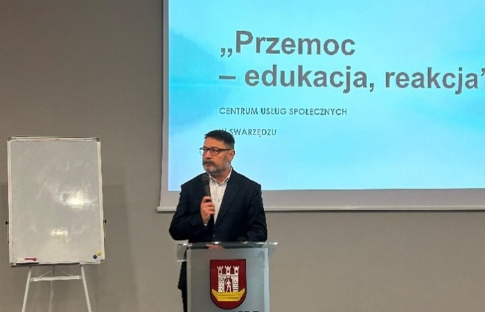 Mężczyzna w garniturze przemawia za mównicą z herbem miasta Swarzędz. Na ekranie wyświetla się temat „Przemoc – edukacja, reakcja”. W tle znajduje się tablica na stojaku