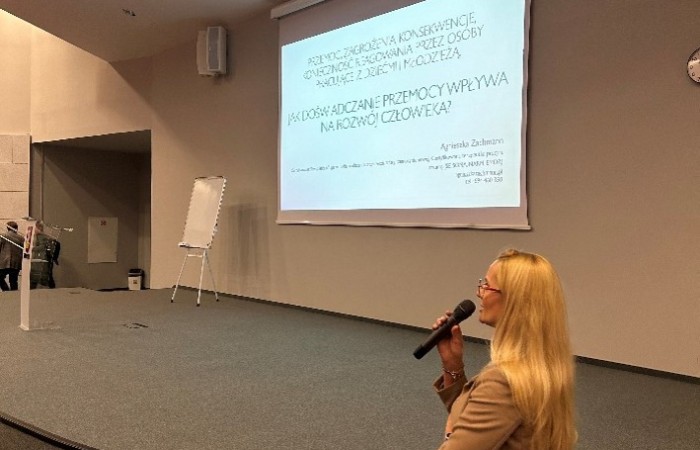 Kobieta o blond włosach, ubrana w beżowy żakiet, przemawia do mikrofonu. W tle znajduje się ekran z wyświetlonym tematem: „Przemoc – zagrożenia i konsekwencje... Jak doświadczanie przemocy wpływa na rozwój człowieka?”. Na lewo od ekranu widać tablicę na stojaku
