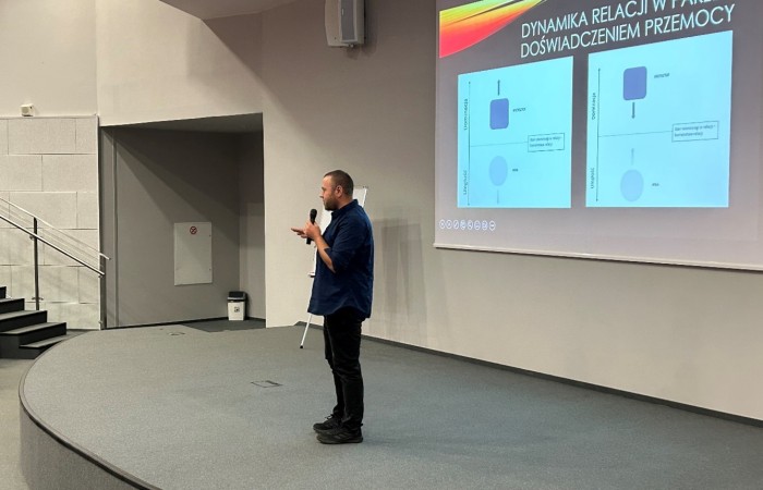 Mężczyzna w granatowej koszuli i czarnych spodniach przemawia na scenie. Na ekranie za nim widoczne są dwa schematy opisujące dynamikę relacji w parze z doświadczeniem przemocy. W tle widać białą ścianę i schody prowadzące na scenę