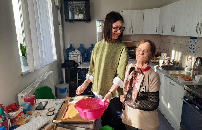 Zdjęcie przedstawia kuchnię w Dziennym Domu Pomocy w Swarzędzu. W centrum fotografii przy stoliku, na którym stoją blachy z ciastem, stoi francuska wolontariuszka Żuljet i uczestniczka zajęć Pani Asia. Żuljet śmieje się i odwraca głowę w stronę Pani Asi. W lewej ręce trzyma łyżkę, a w prawej różową miskę, w której znajduje się masa kremowa. Pani Asia, o głowę niższa od Żuljet, stoi wyprostowana blisko wolontariuszki, ich ramiona stykają się ze sobą. Pani Asia jest oświetlona promieniami słońca wpadającymi przez okno, ma lewą rękę na temblaku, zamknęła oczy uśmiechając się jednocześnie, zupełnie jakby chciała powiedzieć: O, jak bardzo jestem szczęśliwa lub O, jak mi dobrze.