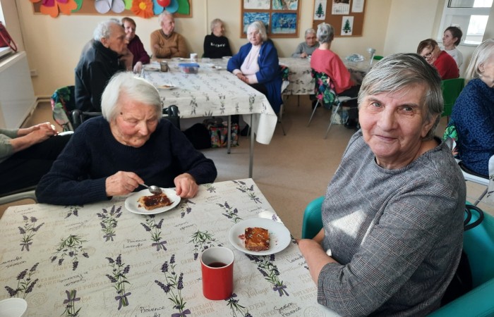 Zdjęcie zostało zrobione z rogu największej sali w Dziennym Domu Pomocy. Przedstawia ono trzynaścioro seniorów siedzących przy stołach przykrytych ceratą z motywami gałązek kwitnącej lawendy. Przed każdą seniorką i każdym seniorem stoi biały talerzyk z kawałkiem ciasta Galet de Rua. Na pierwszym planie widoczna jest uśmiechnięta, siedząca seniorka odwracająca wzrok w kierunku robiącego zdjęcie. Obok niej siedzi inna uczestniczka zajęć, która z zapałem rozpoczyna jedzenie ciasta łyżeczką