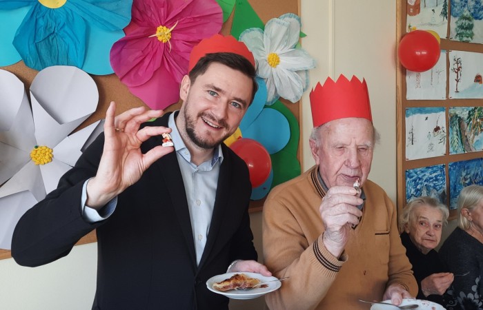 Na tle tablic z pracami plastycznymi wykonanymi przez seniorów z Dziennego Domu Pomocy w Swarzędzu stoi dwóch mężczyzn, którzy mają na głowach korony wykonane z czerwonej tektury. To Dariusz Bugajski pracownik Centrum Usług Społecznych i Antoni Łuczków uczestnik zajęć w Dziennym Domu Pomocy, którzy jedząc Galet de Rua znaleźli w nim porcelanowe figurki fève. Tym samym zostali królami dnia. Jeden i drugi mężczyzna w lewej ręce trzyma talerzyk, a prawą pokazuje swoją figurkę. Za nimi widać siedzącą panią Danusię, która z zaciekawieniem przygląda się figurkom.