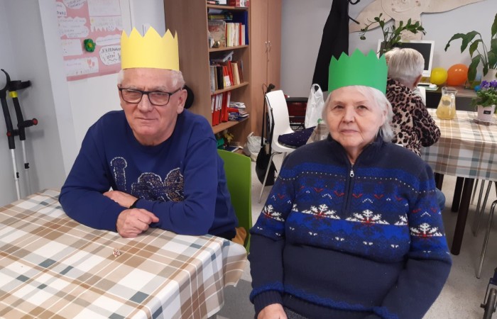 Zdjęcia przedstawia dwie siedzące przy stole postacie, których głowy są przyozdobione kolorowymi koronami. Pani Marylka jest ubrana w granatowy sweter ze świątecznymi wzorami, a Pan Radek w granatową bluzkę z długim rękawem, a na nosie ma czarne okulary. Przed Panem Radkiem na kraciastym obrusie leży mała figurka. Oboje są seniorami uczestniczącymi w zajęciach w Dziennym Domu Pomocy Senior Wigor.