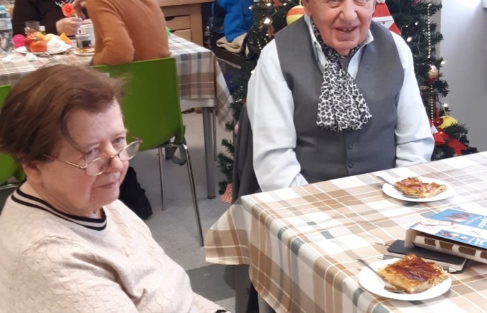 Zdjęcia wykonane zostało w świątecznie udekorowanej sali w Dziennym Domu Pomocy Senior Wigor. Na pierwszym planie widzimy dwie radosne zasiadające przy stole postacie. Po lewej stronie Pani Hania z okularami na nosie, a po prawej elegancko ubrany Pan Mikołaj. Przed Państwem na stole stoją talerzyki z ciastem. W głębi zdjęcia widać sztuczną choinkę ozdobioną bombkami, łańcuchami i światełkami.