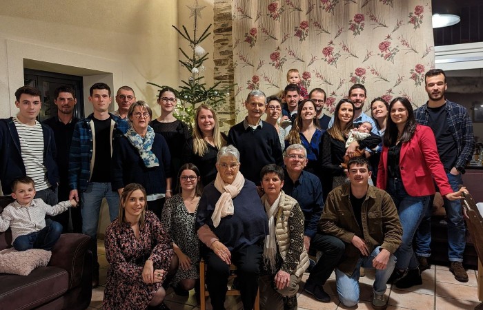 Zdjęcie grupowe przedstawiające rodzinę Żuljet Żama zebraną w salonie domu w czasie Świąt Bożego Narodzenia. Łącznie na zdjęciu widać 26 osób, wśród nich Żuljet. W tle stoi wysoka, ubrana choinka.